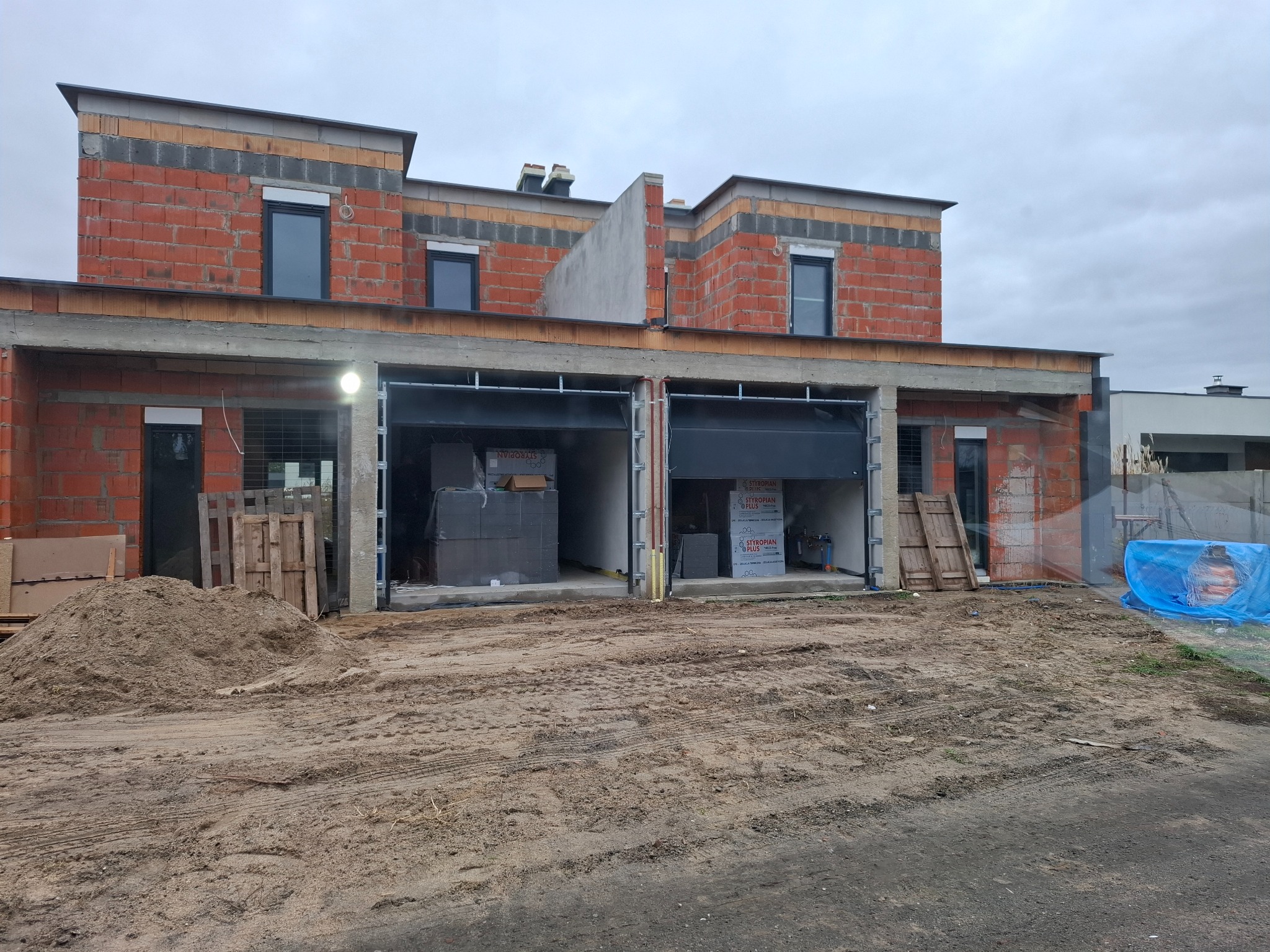 Surowy stan bliźniaka w trakcie budowy: czerwona cegła, szare okna, otwarte garaże z materiałami budowlanymi. Na pierwszym planie piasek i palety.