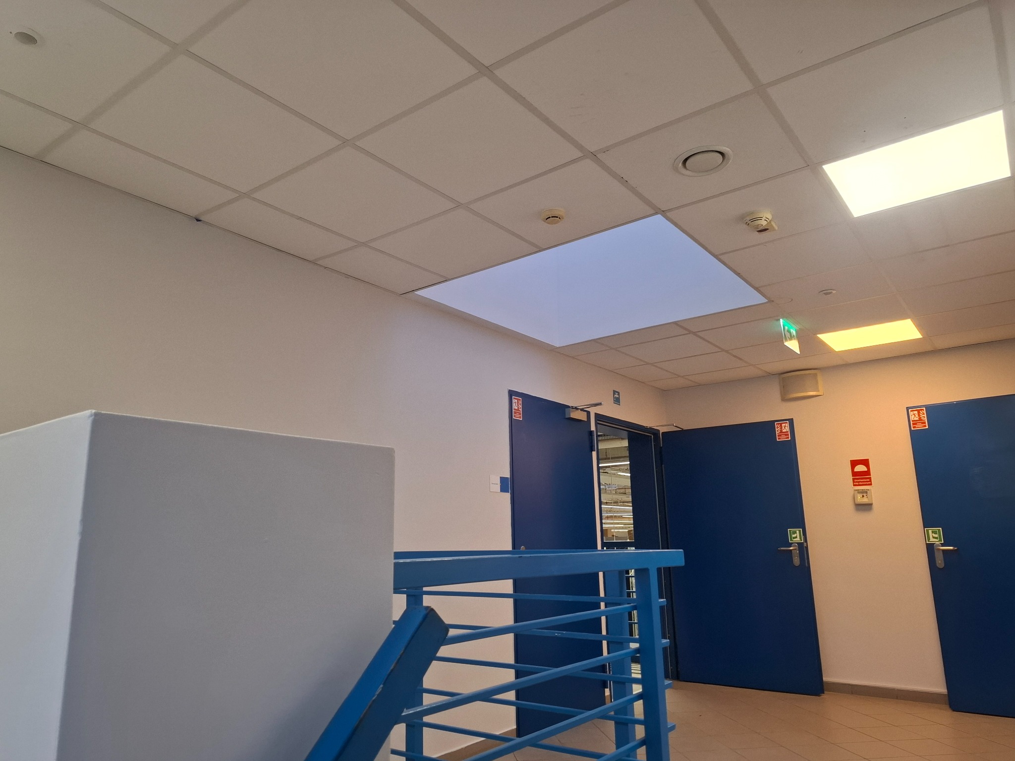 Klatka schodowa z niebieską barierką, białą ścianą i sufitem z panelami oświetleniowymi. Widoczne drzwi w kolorze niebieskim i system przeciwpożarowy.