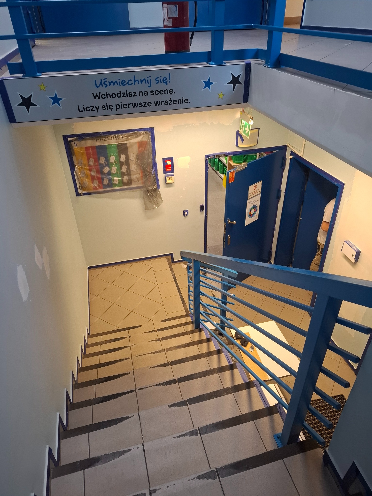 Klatka schodowa w trakcie odświeżania: świeżo pomalowane ściany w jasnym odcieniu, zabezpieczone taśmą schody i framugi drzwi, błękitne barierki i elementy dekoracyjne.