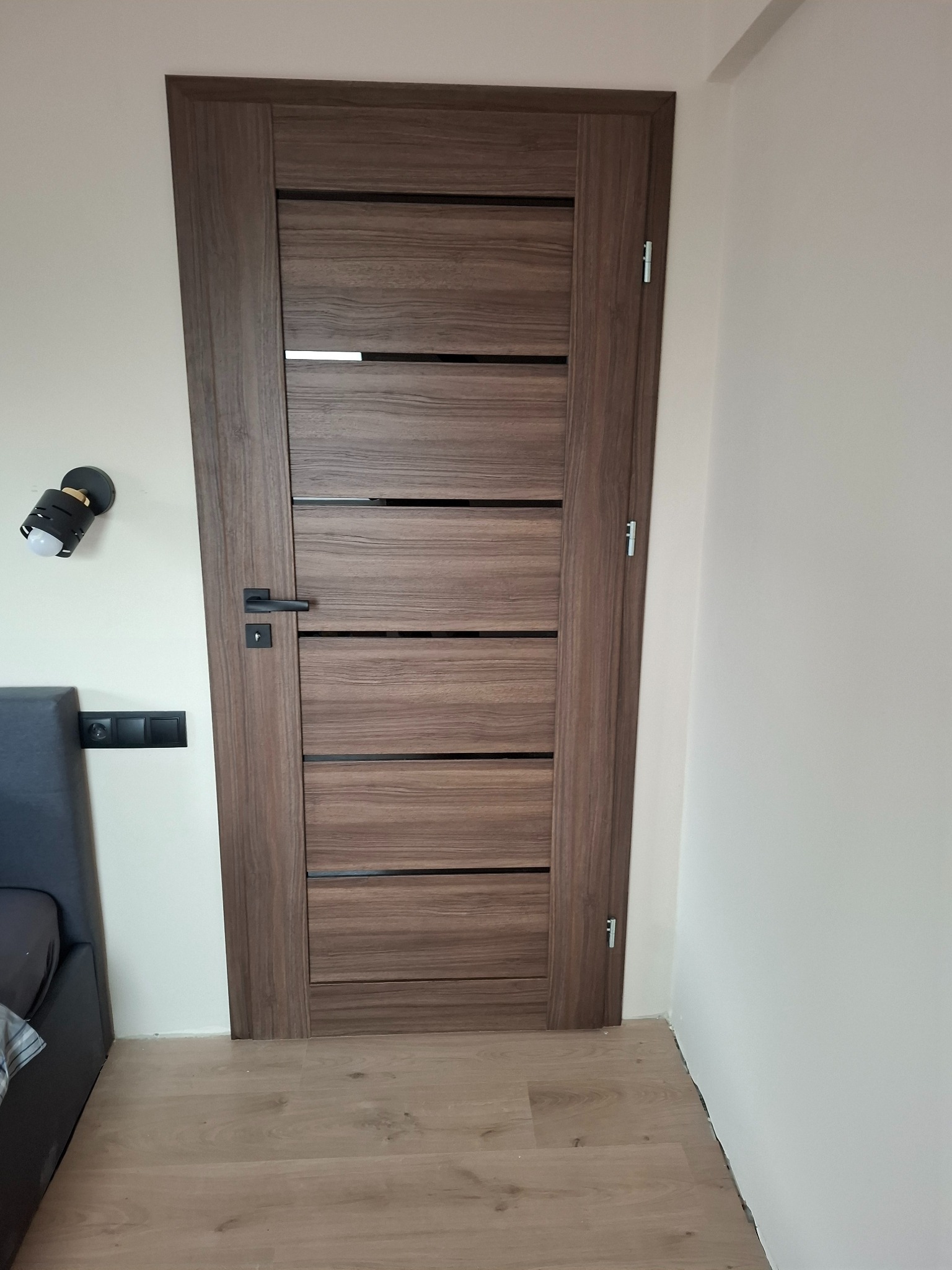 Zamknięte, nowoczesne drzwi wewnętrzne z ciemnego drewna z poziomymi wstawkami, czarną klamką i widocznymi zawiasami, zamontowane w jasnym pomieszczeniu.