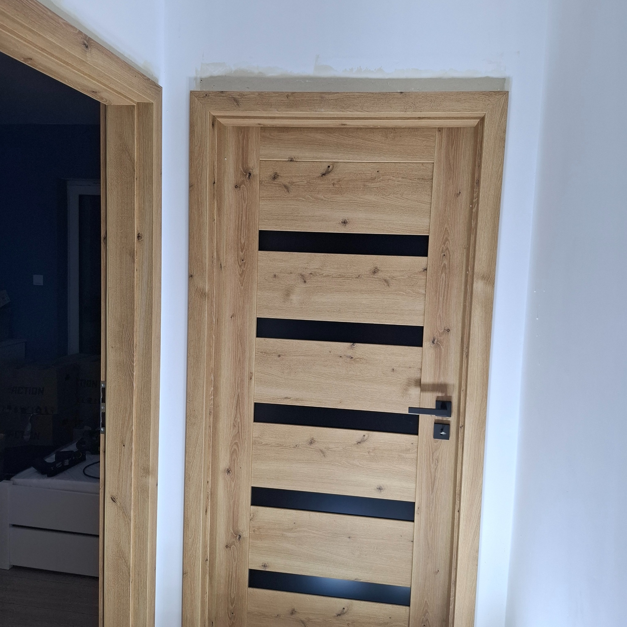 Zamontowane drzwi wewnętrzne z drewnianą okleiną i czarnymi, poziomymi wstawkami, widoczne w nowym, pomalowanym wnętrzu. Minimalistyczny design.