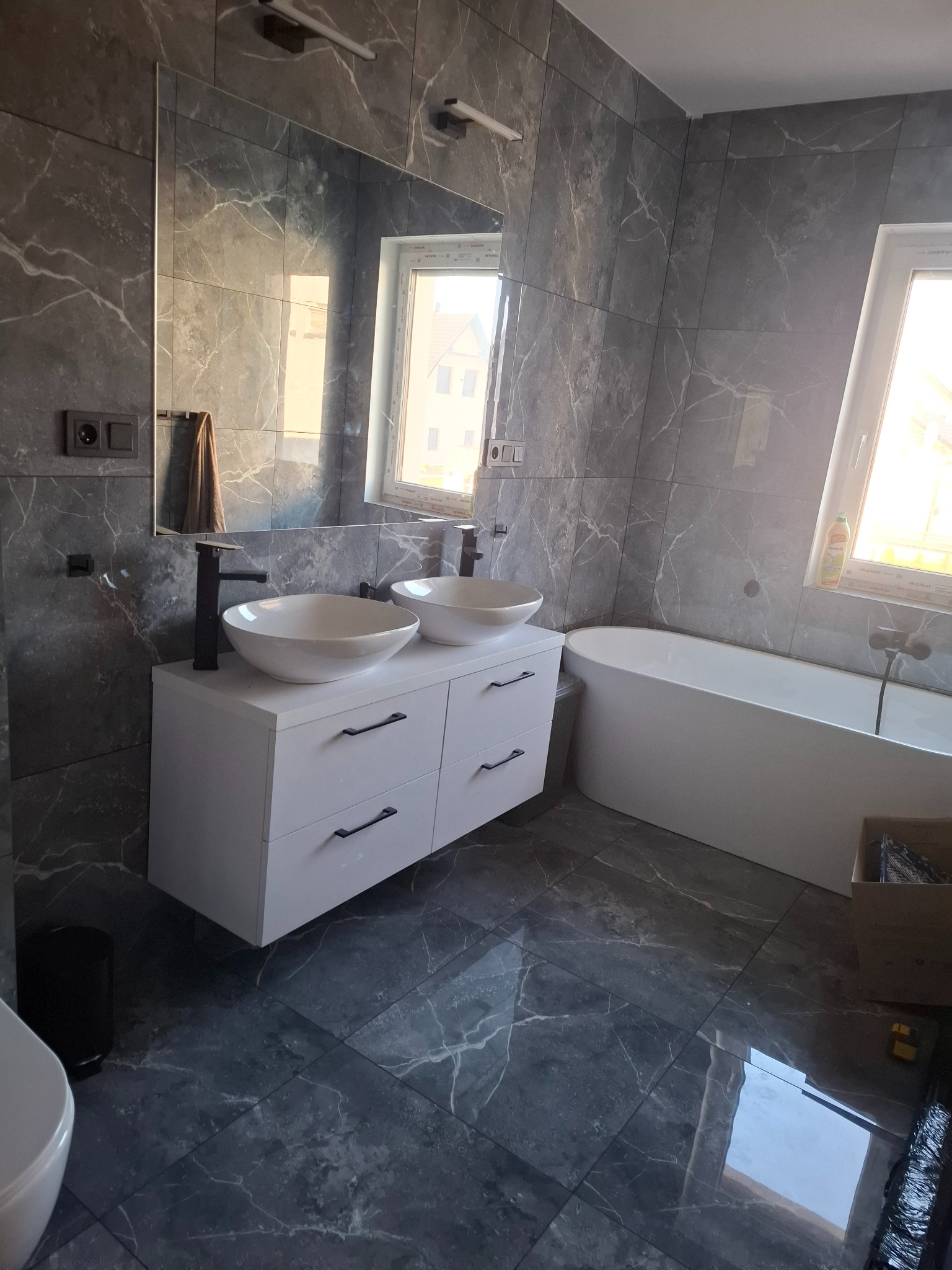 Nowoczesna łazienka z szarymi płytkami imitującymi marmur, białą szafką z dwiema umywalkami i czarną armaturą. Widoczna wanna i okno z widokiem na budynek.