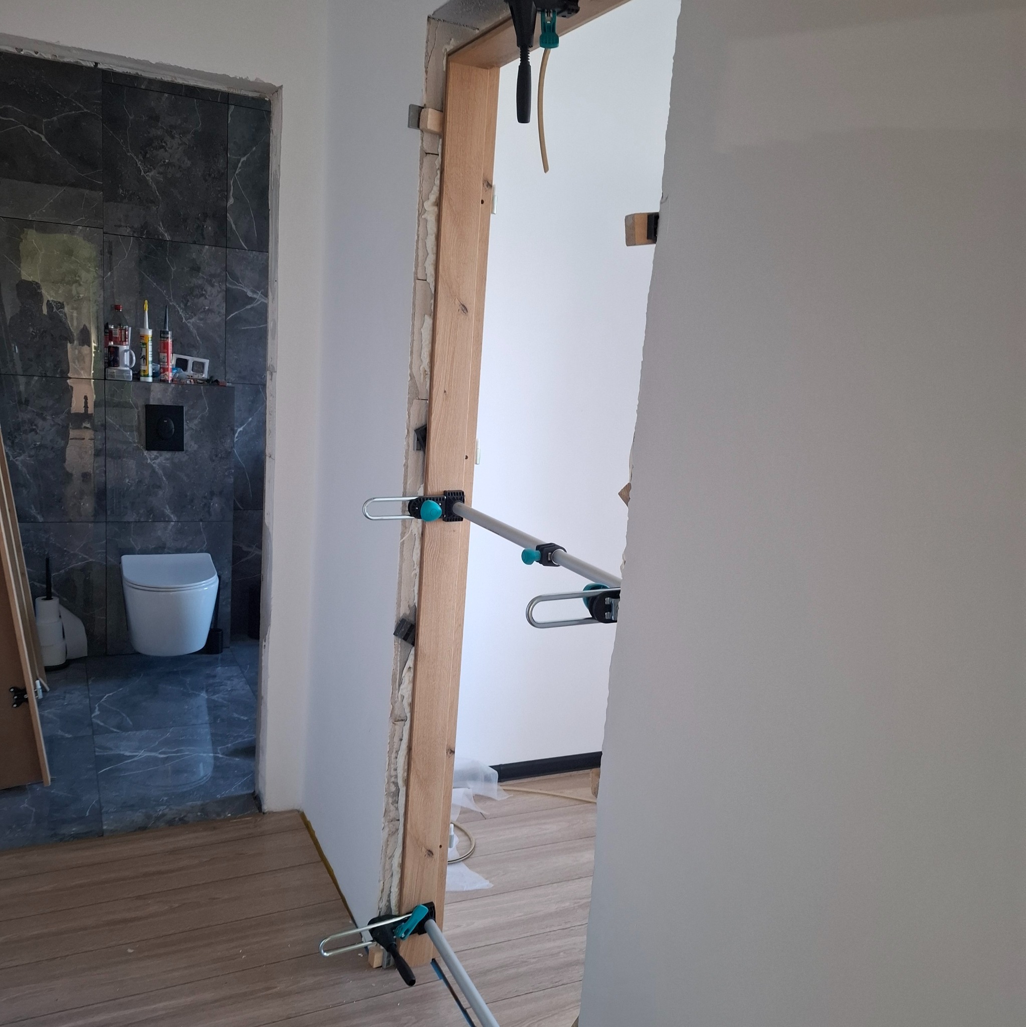 Wnętrze w trakcie remontu: widok na łazienkę z szarymi płytkami imitującymi marmur i toaletą, przez otwór drzwiowy z zamontowaną ościeżnicą i narzędziami.