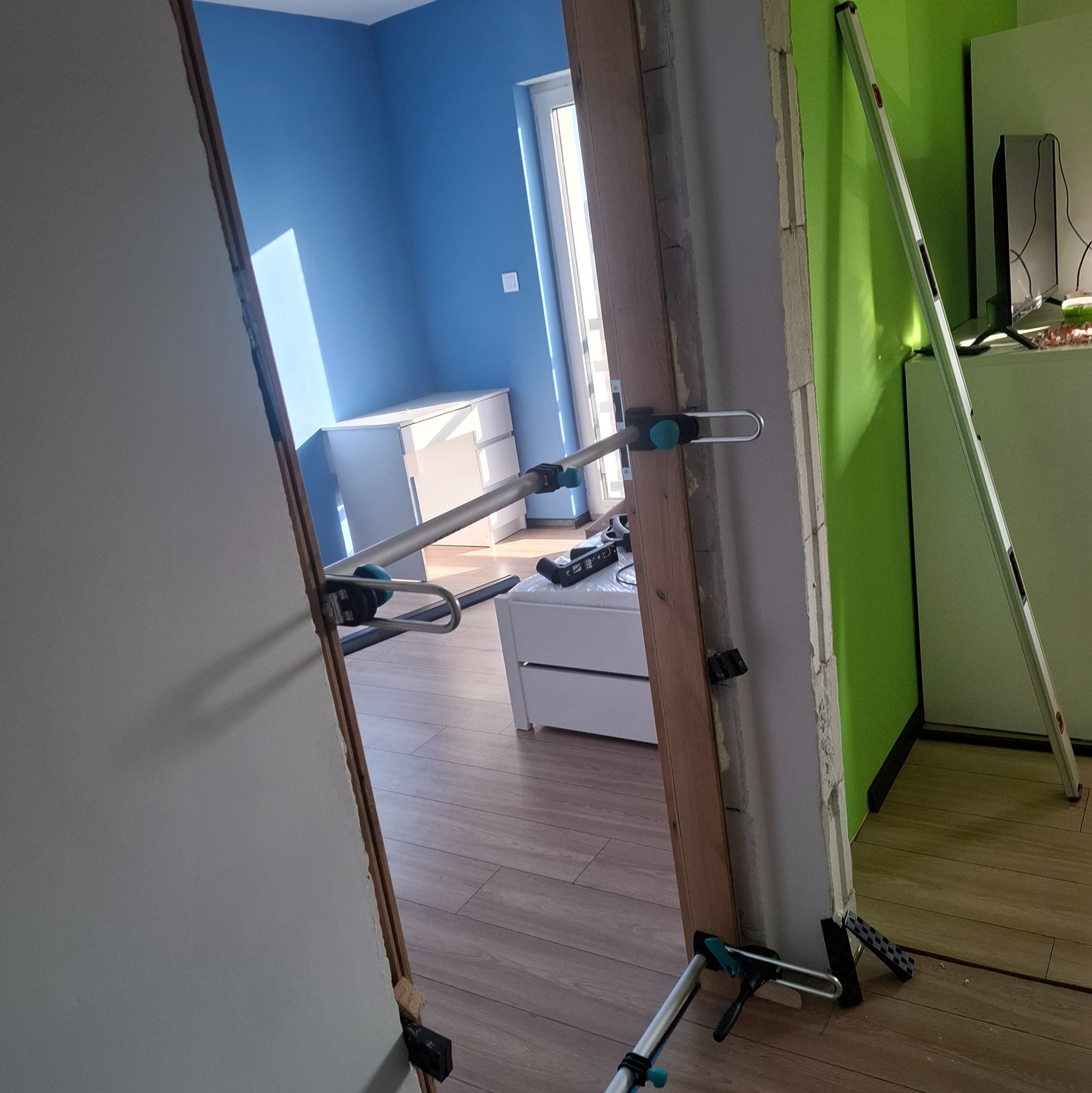 Montaż ościeżnicy drzwiowej z użyciem rozpórki teleskopowej w odnawianym wnętrzu. Widoczne narzędzia i poziomica. Ściany w kolorach niebieskim i zielonym.