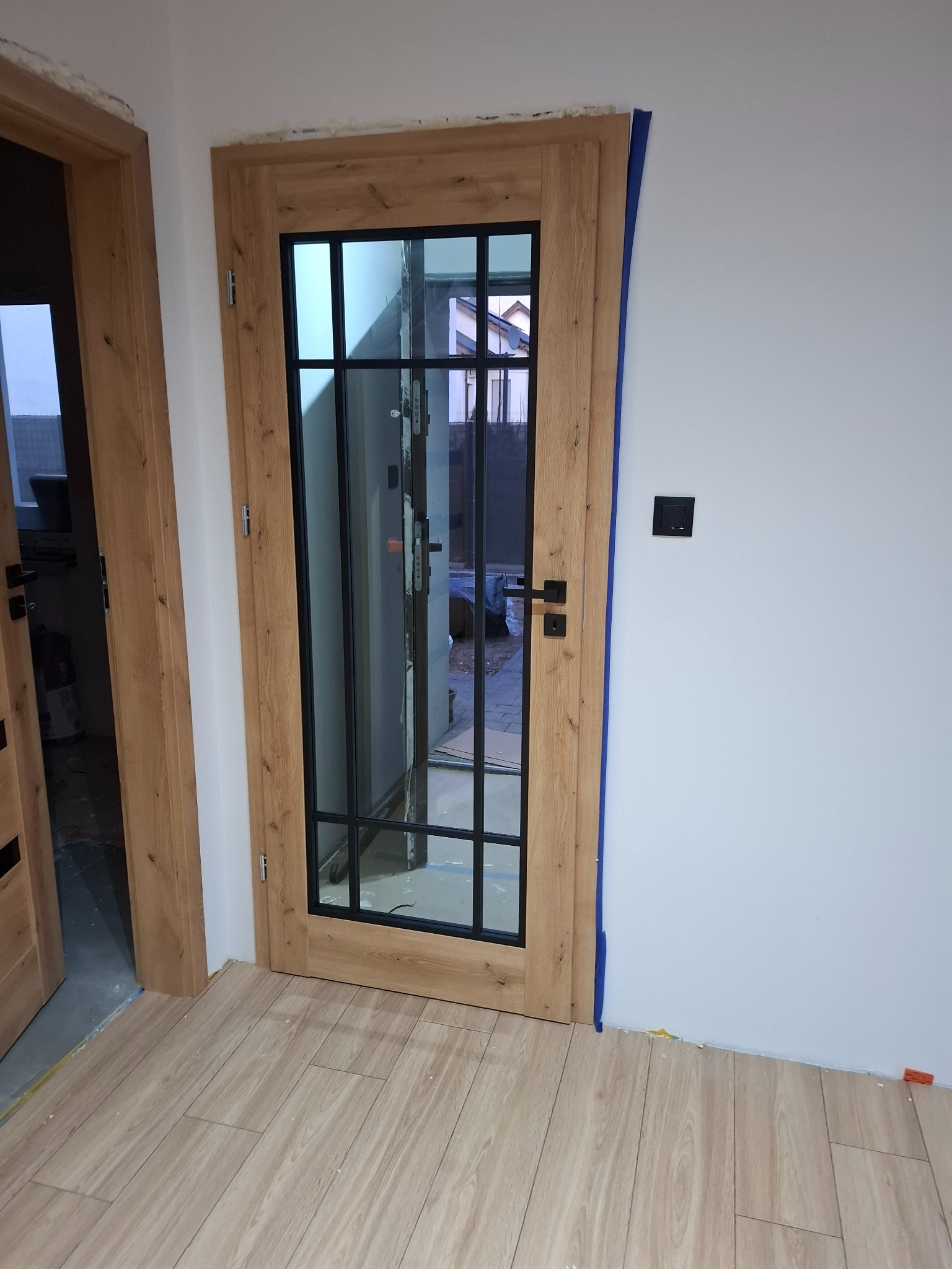 Dwa drewniane, przeszklone drzwi wewnętrzne z czarnymi szprosami i klamkami na tle białej ściany z włącznikiem. Na podłodze panele imitujące drewno.