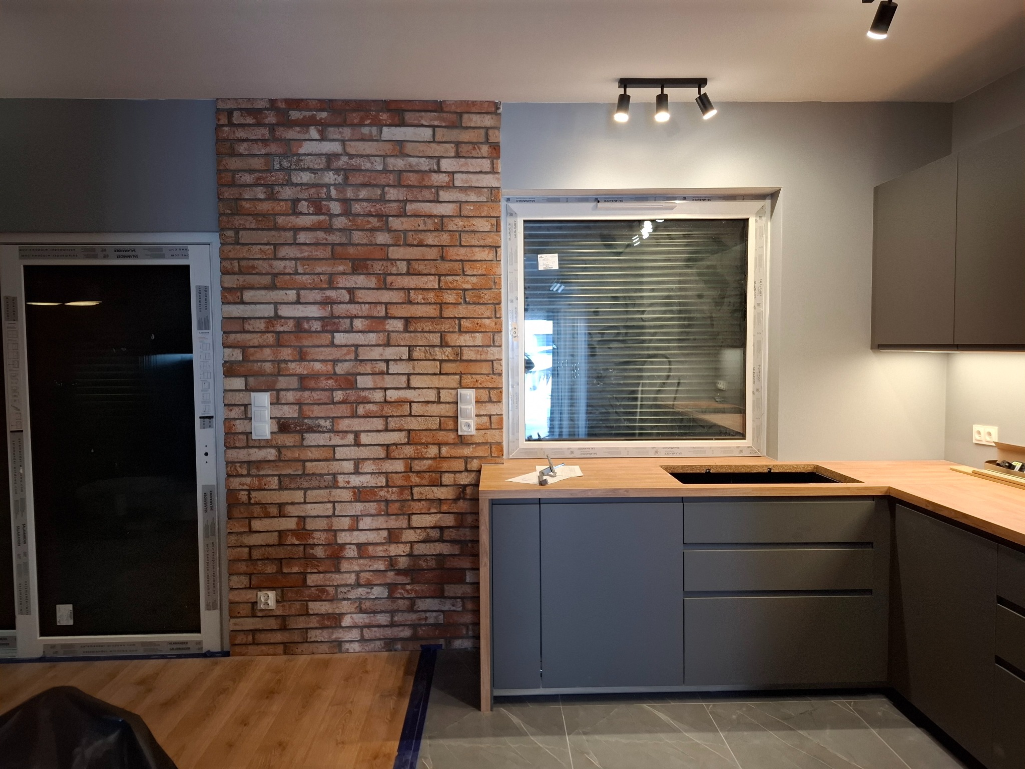 Szara zabudowa kuchenna z drewnianym blatem, ceglana ściana i okno z żaluzjami. Nowoczesne oświetlenie sufitowe. Wykończenie wnętrza w trakcie realizacji.