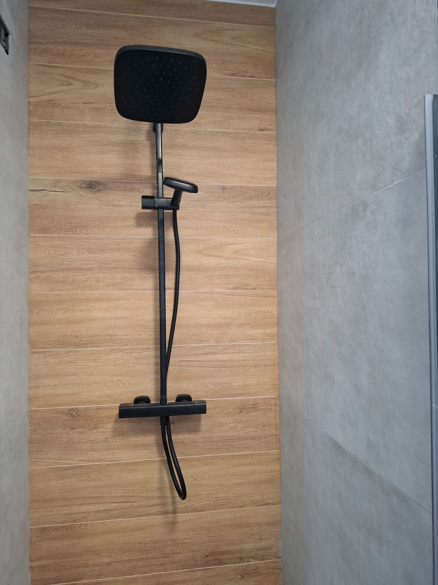 Nowoczesna kabina prysznicowa z czarną armaturą na tle płytek imitujących drewno i szarych, gładkich płytek. Minimalistyczny design, widok z bliska.