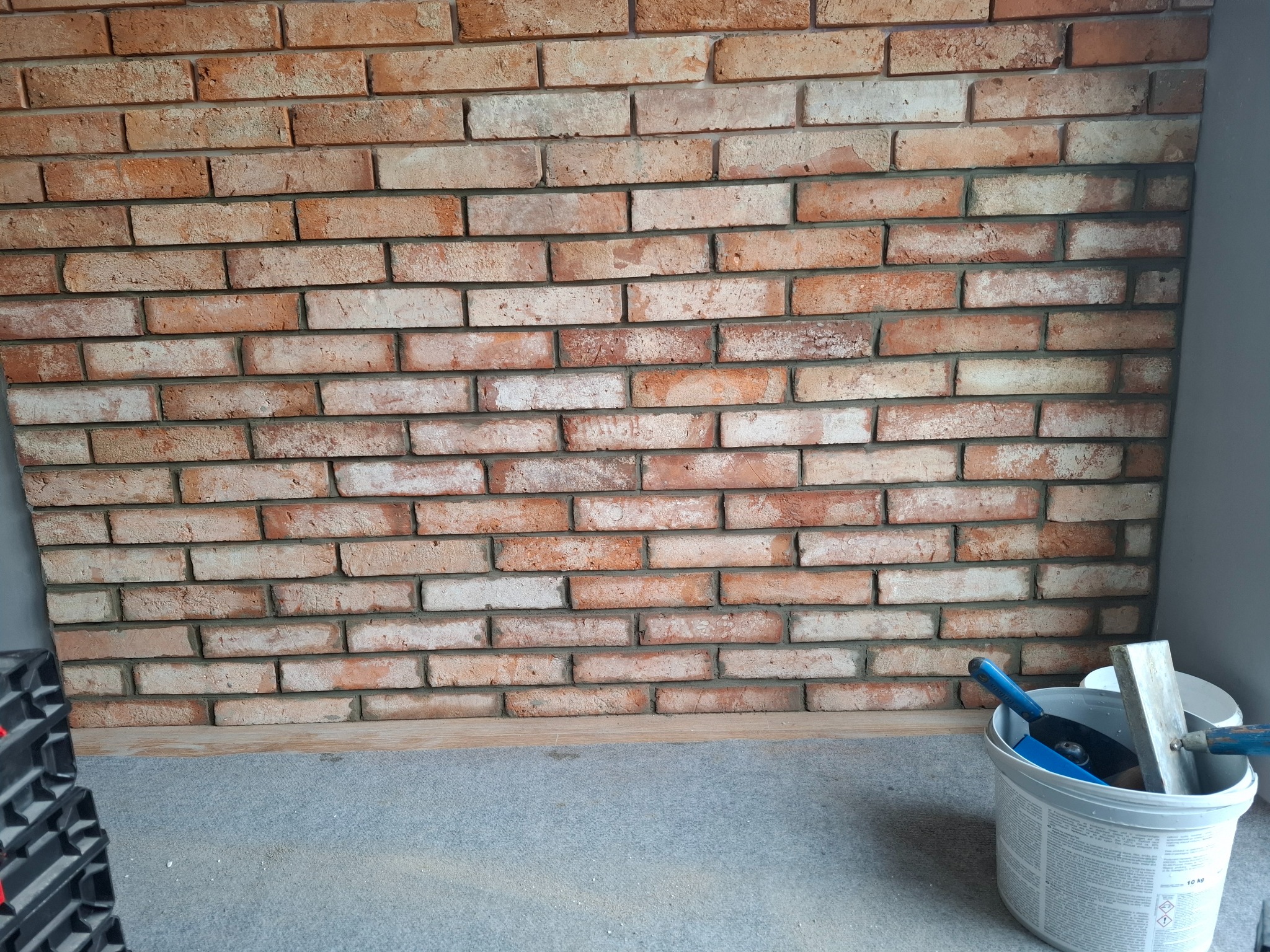 Ściana z cegły klinkierowej w odcieniach czerwieni i brązu, z widocznymi narzędziami glazurniczymi w wiadrze, sugerująca etap prac wykończeniowych.