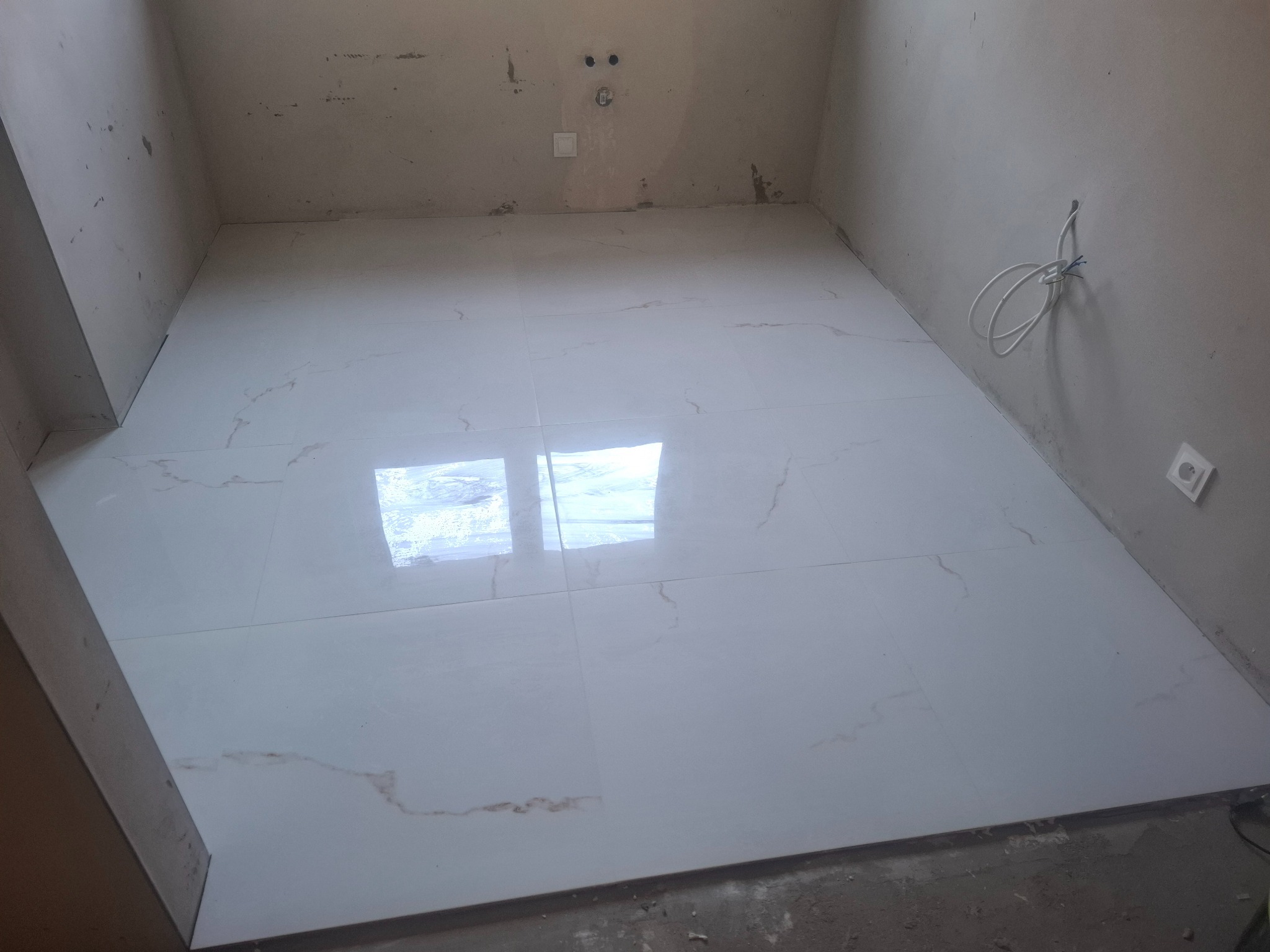 Białe, błyszczące płytki podłogowe z delikatnym wzorem marmuru w surowym wnętrzu łazienki. Widoczne niedokończone ściany i wystające przewody elektryczne.