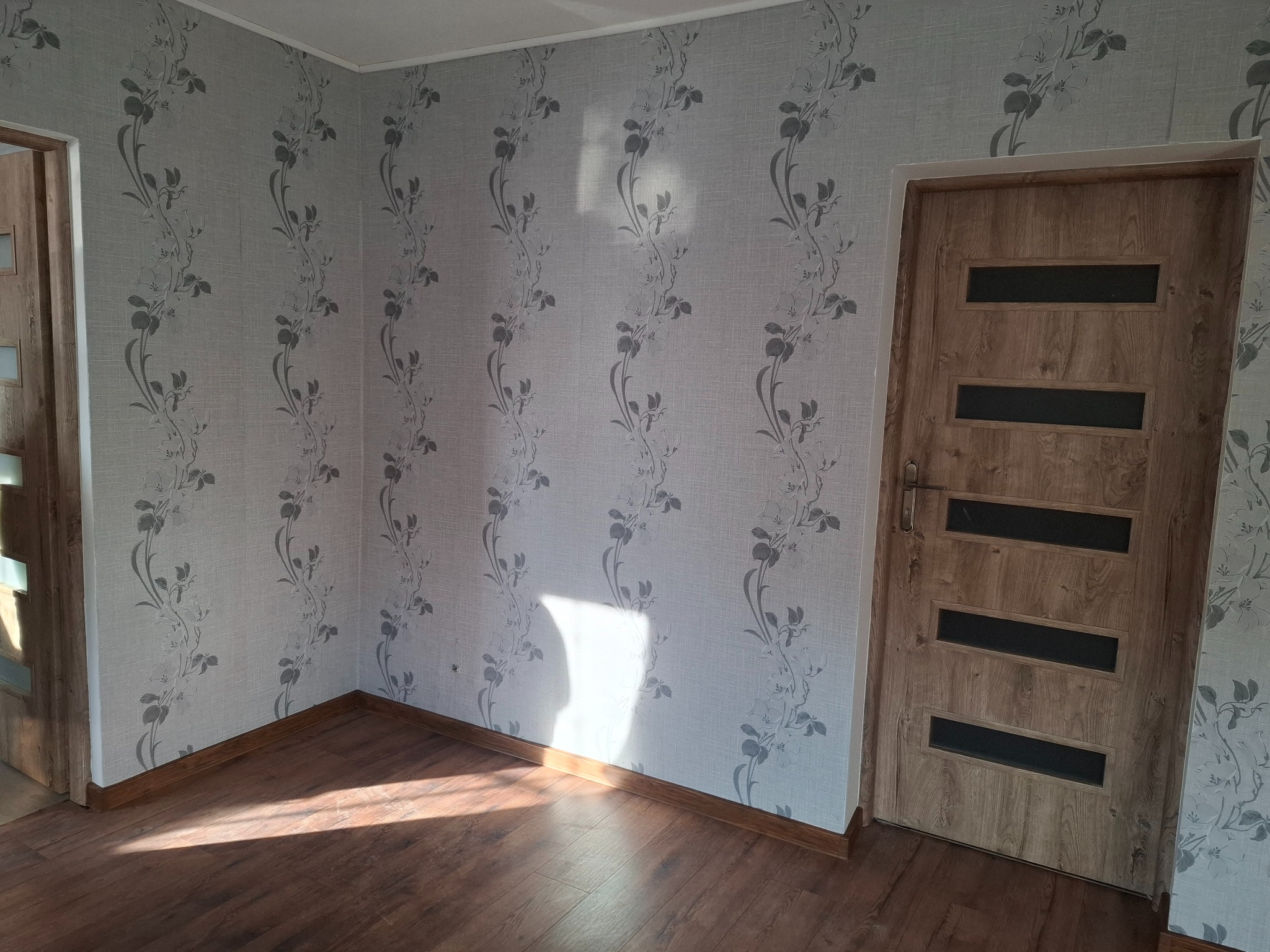 Pokój po remoncie: ściany w jasnej tapecie z motywem roślinnym, drewniana podłoga i drzwi z przeszkleniami. Widoczne listwy przypodłogowe.