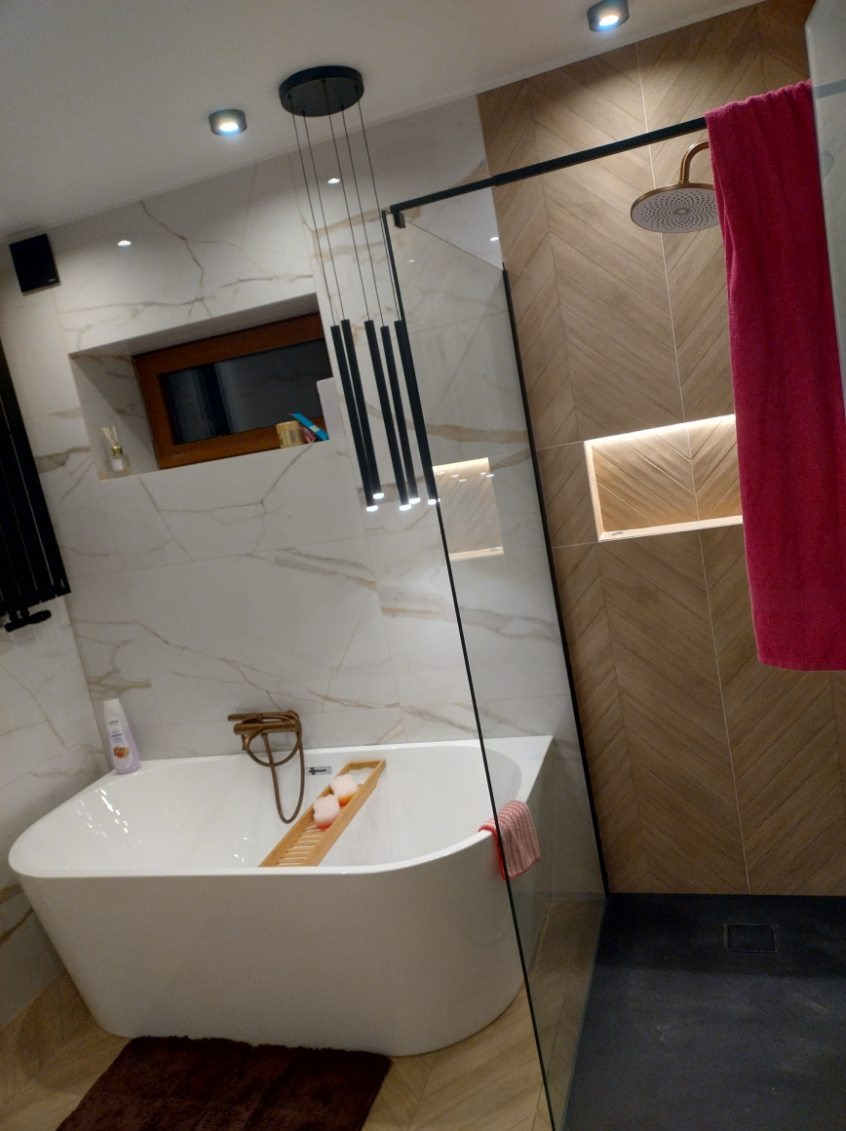 Nowoczesna łazienka z wolnostojącą wanną, marmurowymi płytkami i drewnianymi akcentami. Kabina prysznicowa ze złotą deszczownicą i różowym ręcznikiem.
