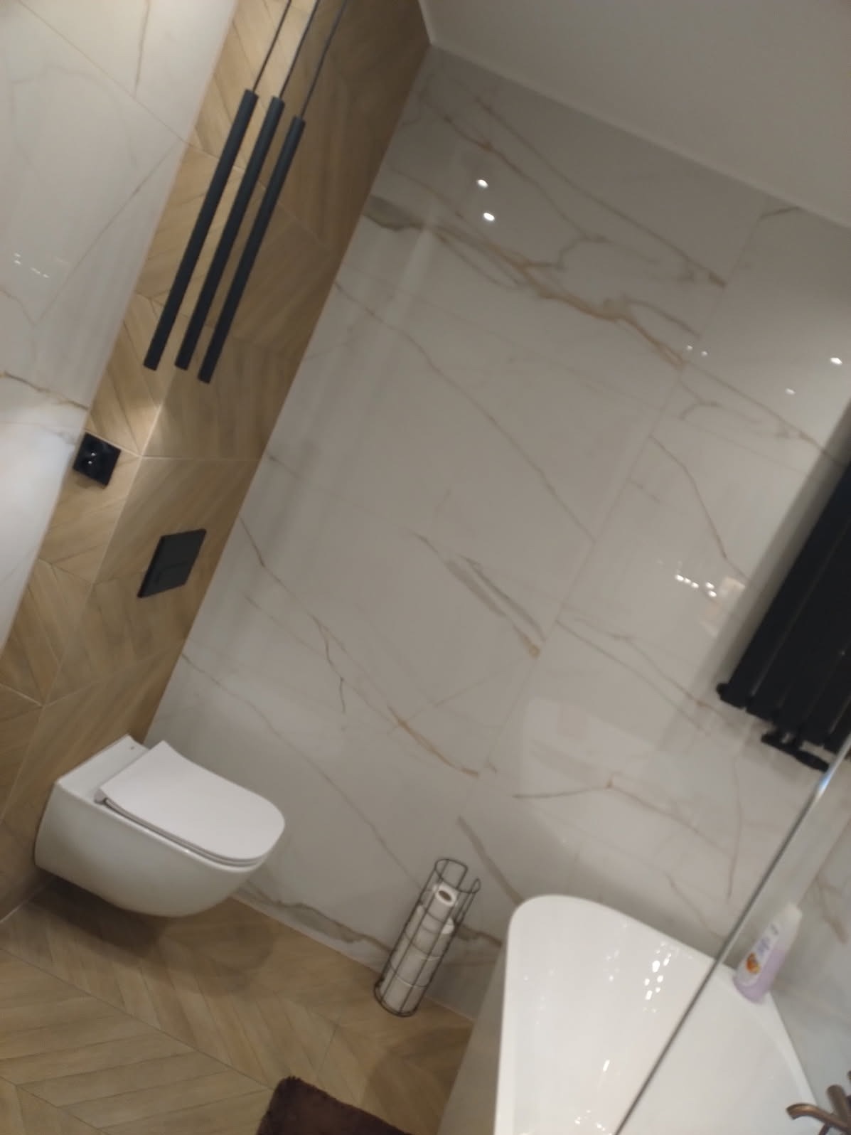 Elegancka łazienka z beżowo-białymi płytkami imitującymi marmur, podwieszana toaleta i czarne, wiszące lampy. Widoczna wanna i grzejnik.