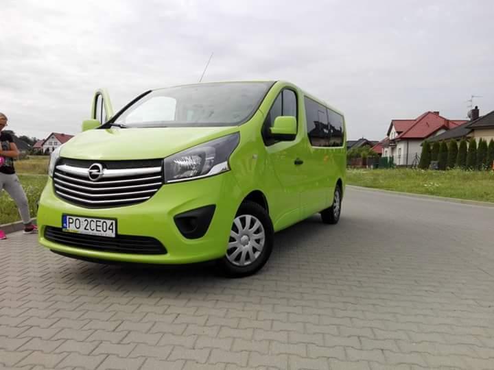 Jasnozielony bus Opel Vivaro z otwartymi drzwiami, zaparkowany na kostce brukowej przed domami jednorodzinnymi.