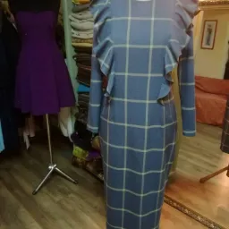 Niebieska sukienka w kratę z falbanami na ramionach, prezentowana na manekinie w pracowni krawieckiej, obok widoczna fioletowa sukienka na drugim manekinie, w tle stosy materiałów.