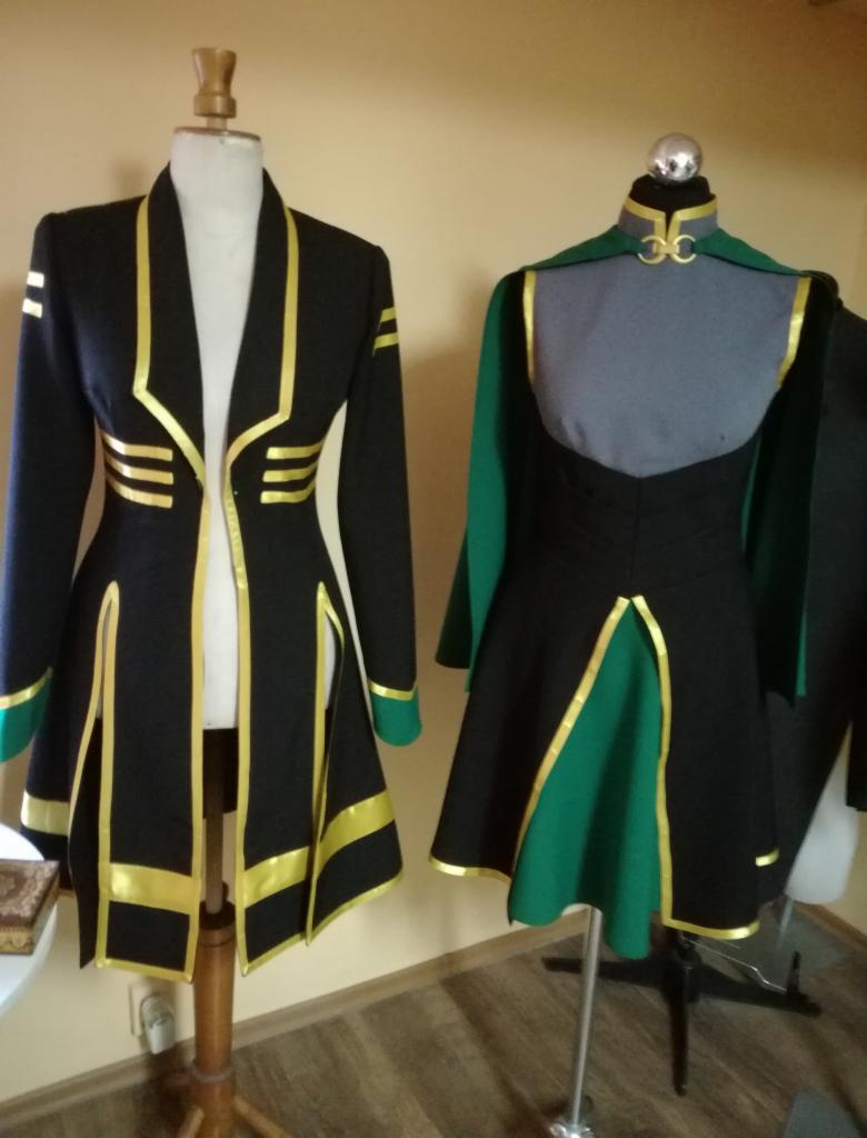 Dwa kostiumy na manekinach: czarny płaszcz z geometrycznymi złotymi zdobieniami i zielonymi mankietami oraz czarno-zielona sukienka z peleryną, również ze złotymi akcentami.