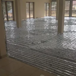 Rozłożone rury ogrzewania podłogowego na siatce w nowym budynku z dużymi oknami, widoczne kolumny podtrzymujące strop.