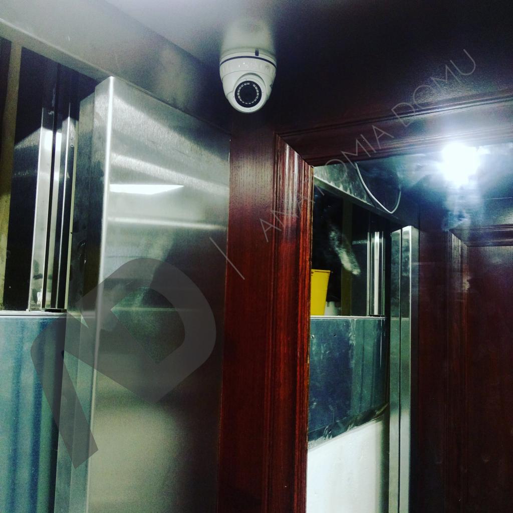 Kamera kopułkowa zamontowana na suficie obok metalowej konstrukcji i drewnianych drzwi, widoczna z dołu pod kątem.