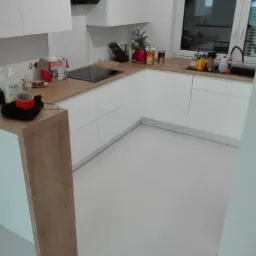 Nowoczesna, biała kuchnia z drewnianym blatem, czarnym okapem i jasną podłogą z płytek, widok na okno z zewnątrz.