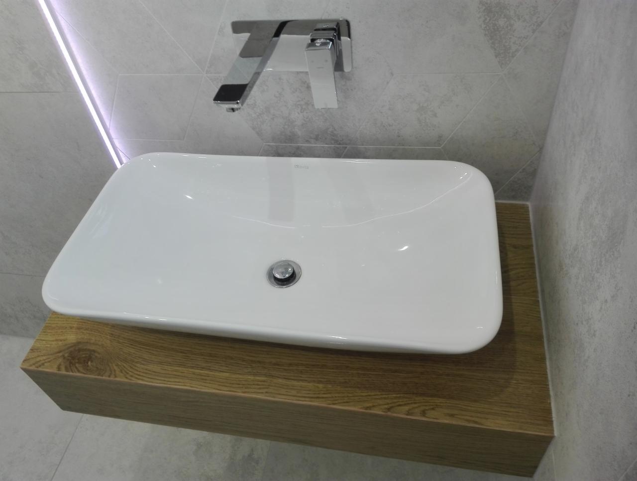 Biała, prostokątna umywalka nablatowa na drewnianej szafce, z chromowaną baterią i szarą, heksagonalną mozaiką na ścianie.