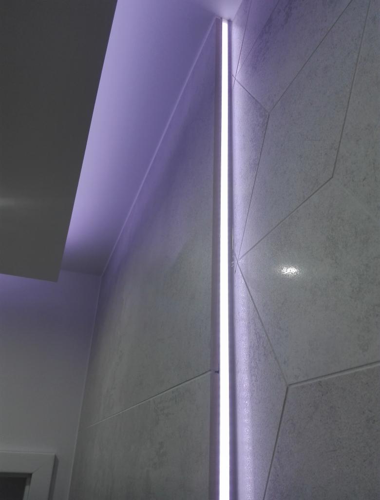 Nowoczesna łazienka z szarymi płytkami o geometrycznym wzorze i pionowym oświetleniem LED wzdłuż krawędzi ściany, tworzącym subtelny, fioletowy blask.