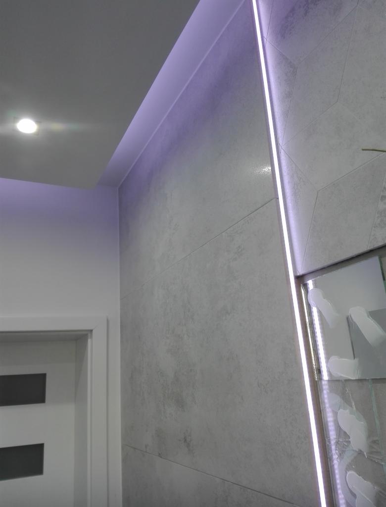 Ściana w łazience wykończona szarymi płytkami imitującymi beton, z podświetleniem LED wzdłuż krawędzi i fragmentem lustra z dekoracyjnymi elementami, oświetlona punktowym światłem.