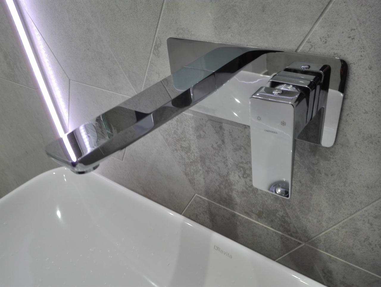 Bateria umywalkowa marki Omnires, zamontowana na szarej, płytkowej ścianie w łazience w Bolesławcu, z widoczną białą umywalką i oświetleniem LED.
