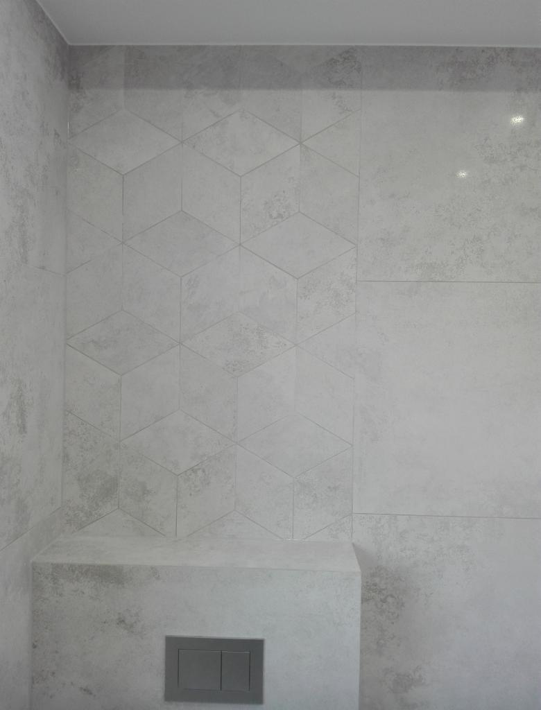 Wykończona ściana w łazience z szarymi płytkami o wzorze geometrycznym 3D, z widocznym zabudowanym stelażem WC z przyciskiem spłukującym.