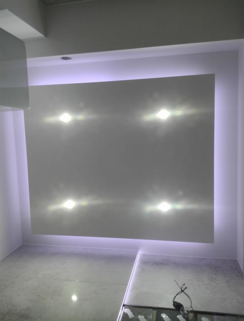 Nowoczesny sufit podwieszany z oświetleniem LED w chłodnej tonacji, cztery punktowe lampy halogenowe, szare płytki podłogowe, fragment zabudowy meblowej widoczny w dolnym rogu.