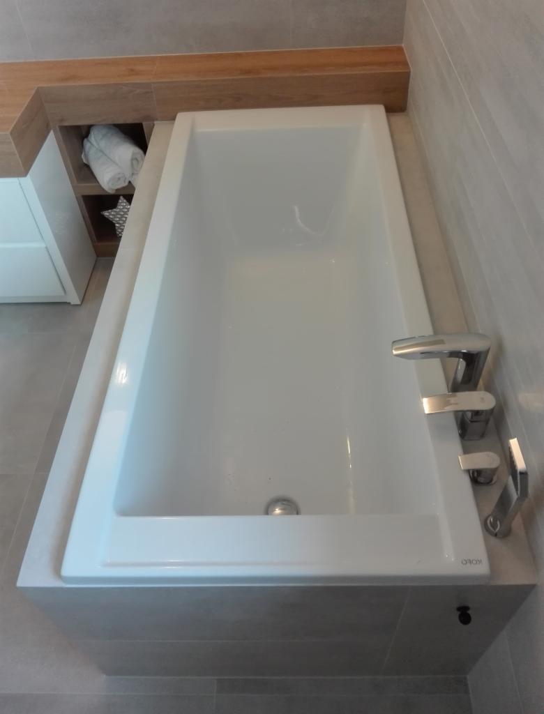 Biała, prostokątna wanna w zabudowie z szarej płytki, bateria chromowana, półka z drewnianym wykończeniem i wnęką na ręczniki w tle.