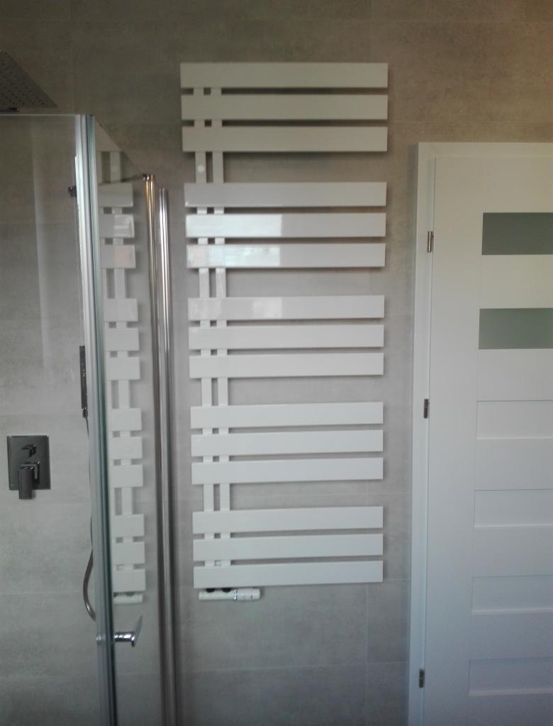 Nowoczesny, biały grzejnik łazienkowy drabinkowy z poziomymi szczeblami, zamontowany na szarej, płytkowej ścianie obok białych drzwi z poziomymi przeszkleniami, widoczny fragment kabiny prysznicowej.