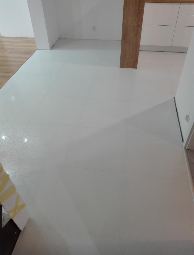 Biała, błyszcząca podłoga z płytek w wyremontowanym pomieszczeniu, widoczne fragmenty ścian i mebli kuchennych, w rogu zabezpieczone folią malarską.