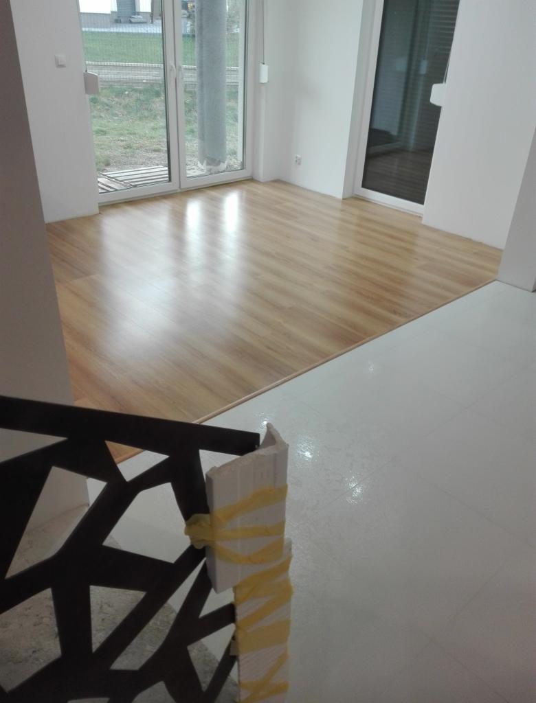 Nowo położona, lśniąca podłoga laminowana w jasnym odcieniu drewna, widoczna z perspektywy schodów z zabezpieczonymi taśmą elementami wykończeniowymi, duże okna balkonowe w tle.