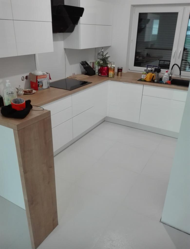 Nowoczesna, biała kuchnia z drewnianym blatem, czarnym okapem i jasną podłogą z płytek, widok na okno z zewnątrz.