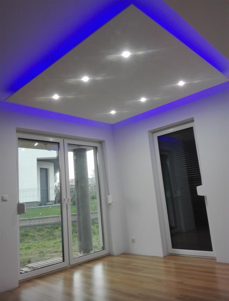 Nowoczesne wnętrze: białe ściany, drewniana podłoga, sufit podwieszany z oświetleniem LED w kolorze niebieskim i punktowymi lampami, widok na zewnątrz przez duże okna.