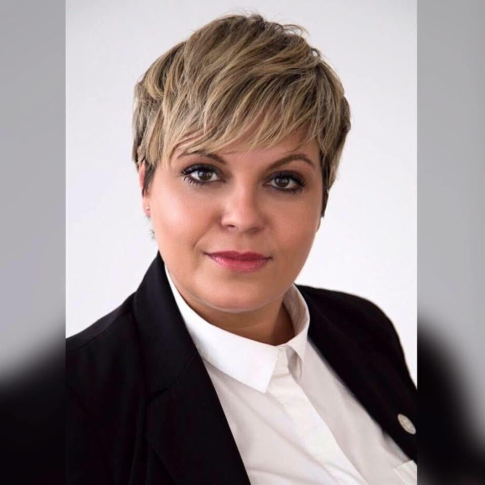 Portret kobiety w czarnej marynarce i białej koszuli, z krótkimi blond włosami i subtelnym makijażem, uśmiechająca się delikatnie, na białym tle.