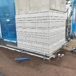 Kładzenie szarych płytek elewacyjnych na rogu budynku, widoczne krzyżyki dystansowe, fragment rusztowania, ziemia i niebieski wąż.