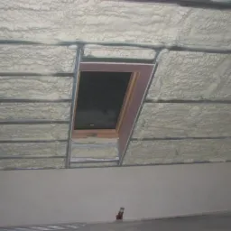 Poddasze z oknem dachowym w trakcie izolacji pianą poliuretanową, widoczne metalowe profile konstrukcyjne i fragment ściany szczytowej.