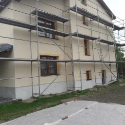 Dwukondygnacyjny dom z jasną elewacją w trakcie prac wykończeniowych, otoczony metalowym rusztowaniem; teren wokół zabezpieczony białą włókniną.