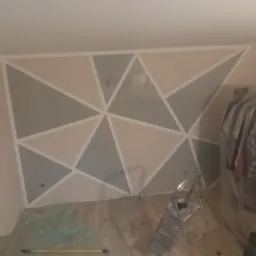 Ściana w pokoju na poddaszu z geometrycznym wzorem malowanym w odcieniach szarości i beżu, zabezpieczona folią podłoga, widoczna drabina malarska i ubrania na wieszakach.