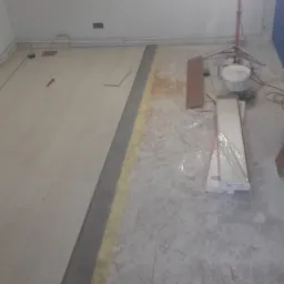 Układanie jasnych paneli podłogowych, widoczny fragment ułożonej podłogi, listwa dylatacyjna, paczka paneli w folii, wiadro z klejem i lampa budowlana w tle.