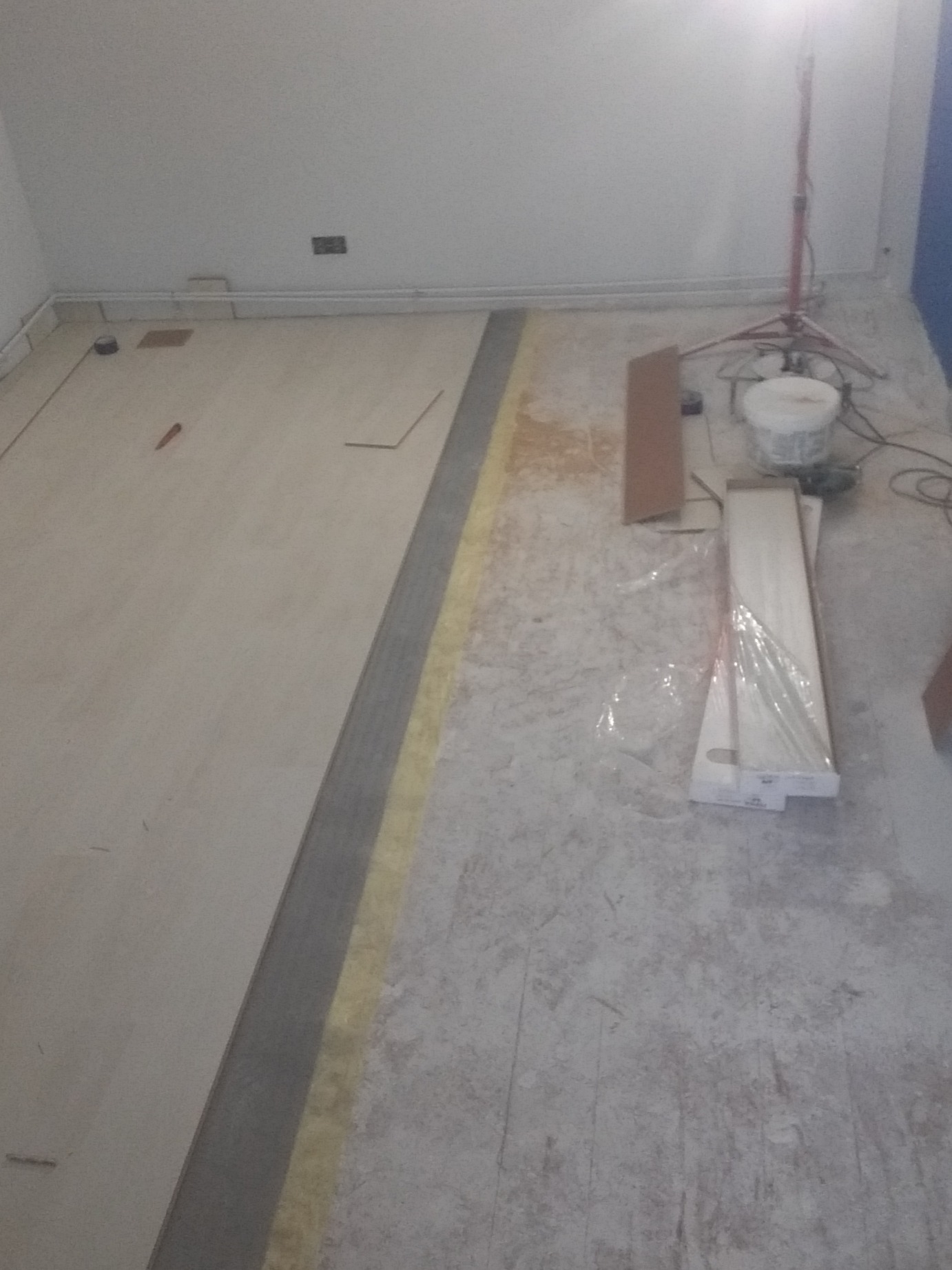 Układanie jasnych paneli podłogowych, widoczny fragment ułożonej podłogi, listwa dylatacyjna, paczka paneli w folii, wiadro z klejem i lampa budowlana w tle.