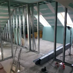 Montaż metalowej konstrukcji pod płyty karton-gips na poddaszu ze skosami i oknami dachowymi, widoczne profile, częściowo wykończone ściany i sufit, drabina i lampa budowlana.