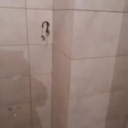 Ściana wyłożona beżowymi płytkami ceramicznymi, widoczne otwory na rury i wystające przewody elektryczne.