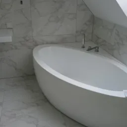 Nowoczesna łazienka z wolnostojącą wanną o owalnym kształcie, marmurowe płytki na ścianach i podłodze, minimalistyczna umywalka wisząca, sufit ze skosem.