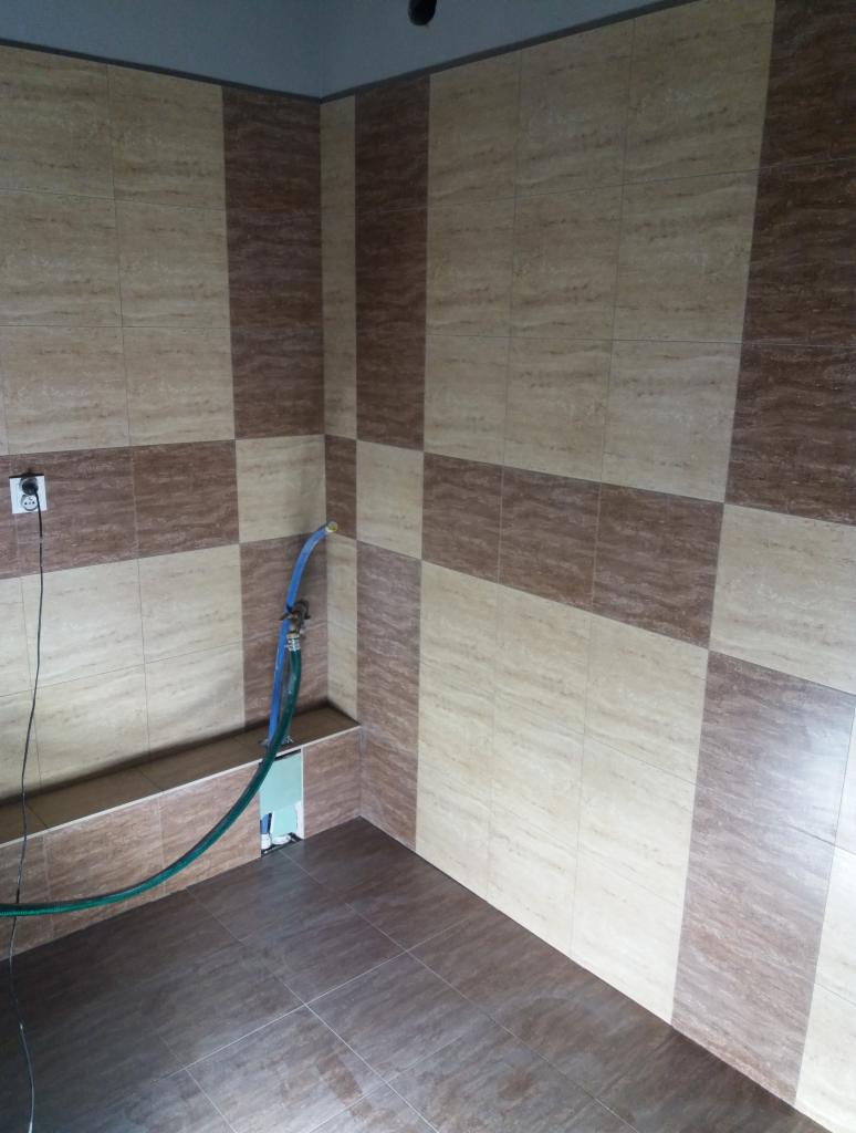 Łazienka w trakcie wykańczania z ułożonymi płytkami w odcieniach beżu i brązu, widoczny wąż doprowadzający wodę i gniazdko elektryczne.