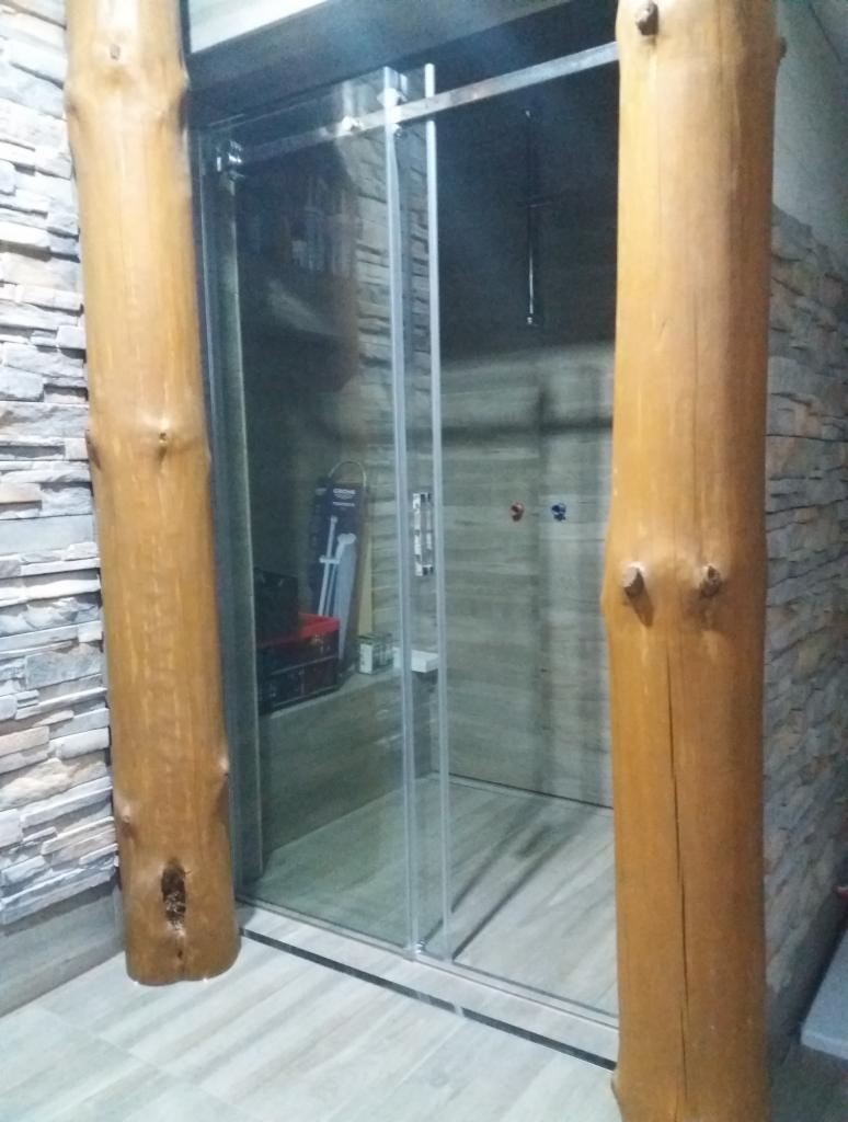 Szklana kabina prysznicowa z przesuwnymi drzwiami, otoczona dekoracyjnymi słupami z naturalnego drewna, na tle ściany wyłożonej kamieniem w odcieniach szarości.