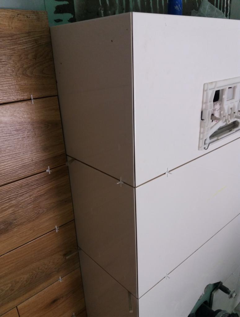 Układanie płytek imitujących drewno na ścianie obok zabudowy stelaża WC z białych płytek, widoczne krzyżyki dystansowe.