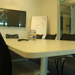 Property Investment sp zo.o. - Sala konferencyjna z białym stołem, krzesłami z siatkowym oparciem, telewizorem na ścianie i flipchartem z wykresami, butelkami wody i soków na stole.