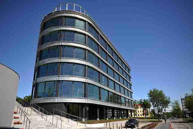 Nowoczesny, zaokrąglony budynek biurowy z dużą ilością okien, widoczny z poziomu ulicy, z schodami prowadzącymi do wejścia i zaparkowanym samochodem w pobliżu.