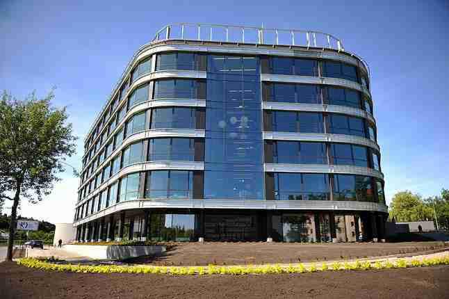 Nowoczesny, sześciopiętrowy budynek biurowy z zaokrąglonymi narożnikami i szklaną fasadą, widziany z poziomu gruntu w słoneczny dzień.